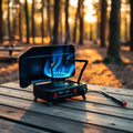 Black cassette gas stove with blue flame on wooden camp table at sunset