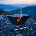 V-shaped folding charcoal BBQ grill with glowing embers at dusk on rocky terrain
